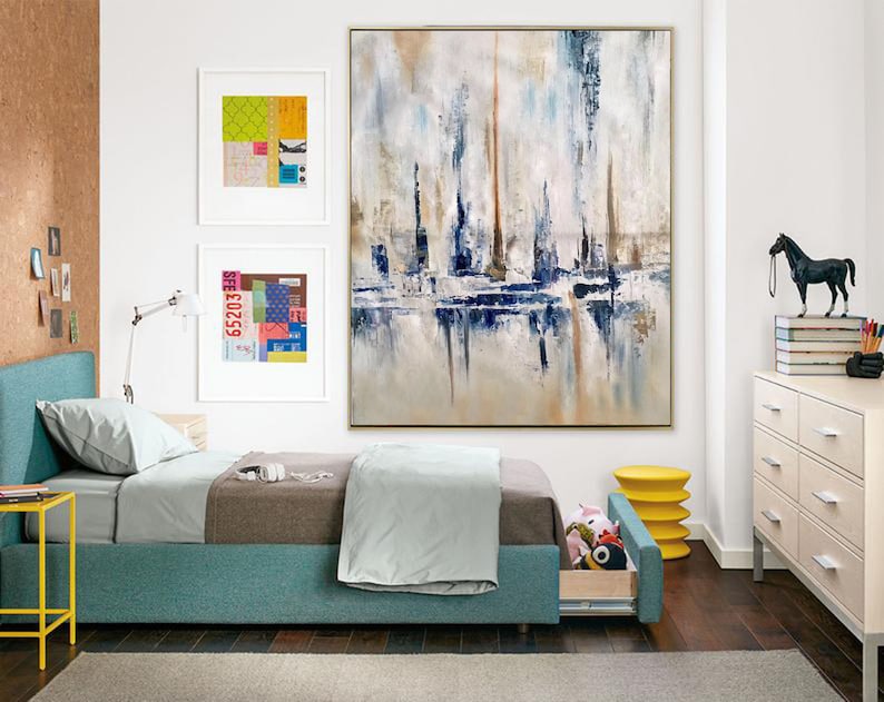 May include: An abstract painting in a gold frame, featuring vertical brushstrokes in white, blue, and brown. The artwork hangs above a bed with a teal frame and a gray comforter. Two framed collages and a yellow side table are also visible.