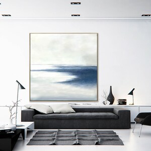 May include: Large abstract painting with a gold frame, featuring a serene seascape in shades of blue and white. The artwork hangs above a gray sofa, complementing the modern living room decor. The room includes a black rug, lamps, and minimalist furniture.