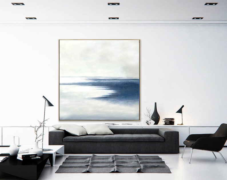 May include: Large abstract painting with a gold frame, featuring a serene seascape in shades of blue and white. The artwork hangs above a gray sofa, complementing the modern living room decor. The room includes a black rug, lamps, and minimalist furniture.