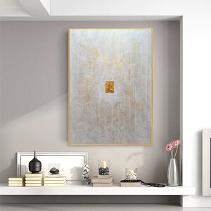 May include: A rectangular wall art piece featuring a textured white surface with intricate, wavy gold lines and a central, solid gold rectangular accent. The artwork is enclosed in a thin gold frame, creating a visual focal point with its layered, embossed appearance and metallic colour palette.