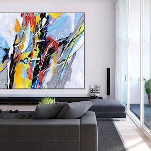 May include: Abstract painting with bold strokes of yellow, blue, red, black, and white on a gray background. The painting is hanging on a white wall in a modern living room.