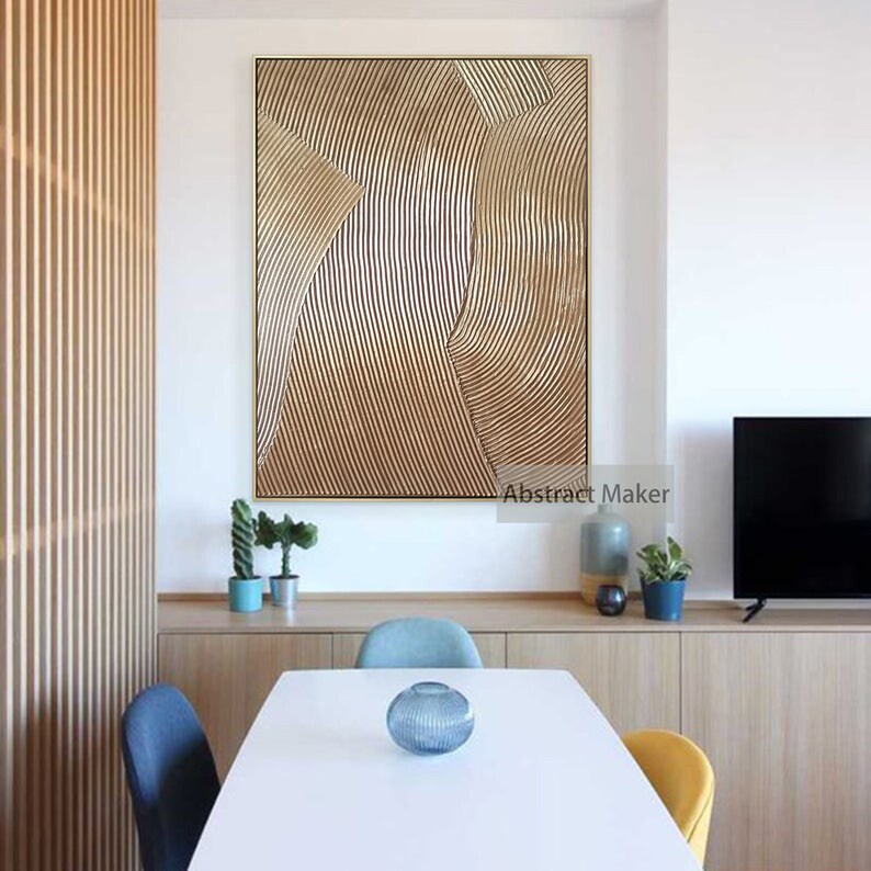 Puede incluir: Pintura abstracta con l&iacute;neas y curvas doradas y marrones. La pintura est&aacute; enmarcada en oro y cuelga de una pared blanca sobre una mesa de madera con sillas.