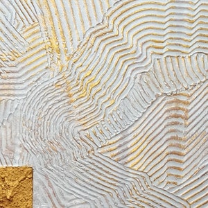 May include: A textured wall art piece featuring a white surface with raised, parallel lines in a wavy pattern. The ridges are accented with metallic gold paint, and a solid, square-shaped block with a rough, gold-painted texture sits in the corner.