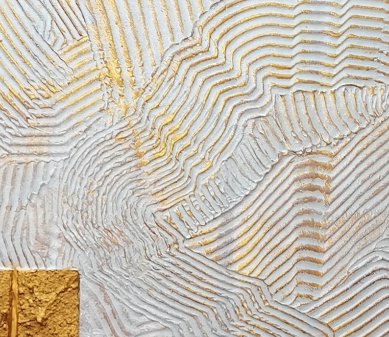 May include: A textured wall art piece featuring a white surface with raised, parallel lines in a wavy pattern. The ridges are accented with metallic gold paint, and a solid, square-shaped block with a rough, gold-painted texture sits in the corner.