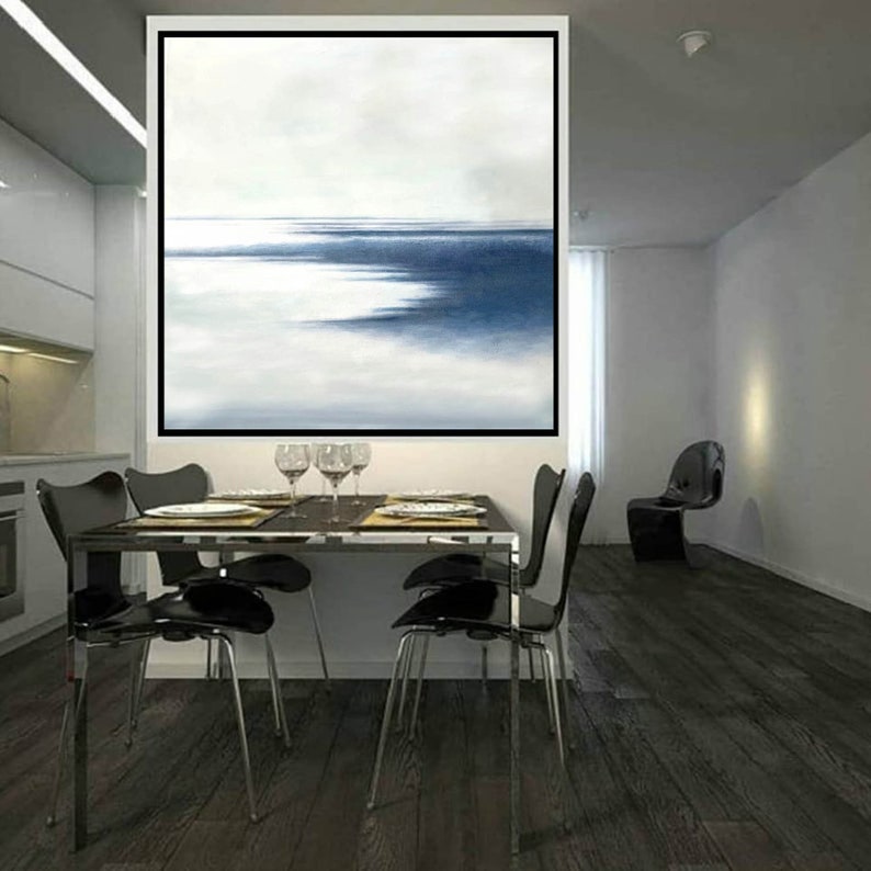 May include: An abstract painting with a black frame depicts a seascape in shades of blue and white. The artwork hangs above a dining table set with plates, glasses, and four black chairs. The room has dark wood flooring and a modern aesthetic.