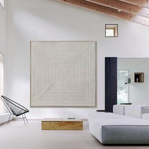 May include: A white, square, abstract painting with a geometric pattern of lines. The painting is hanging on a white wall in a modern living room with a white couch and a black chair.