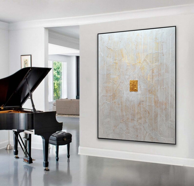 May include: A large rectangular wall art piece featuring a white textured surface with wavy, embossed lines and gold accents. A central, raised gold rectangular element is positioned in the middle of the canvas, which is framed in a thin black border.