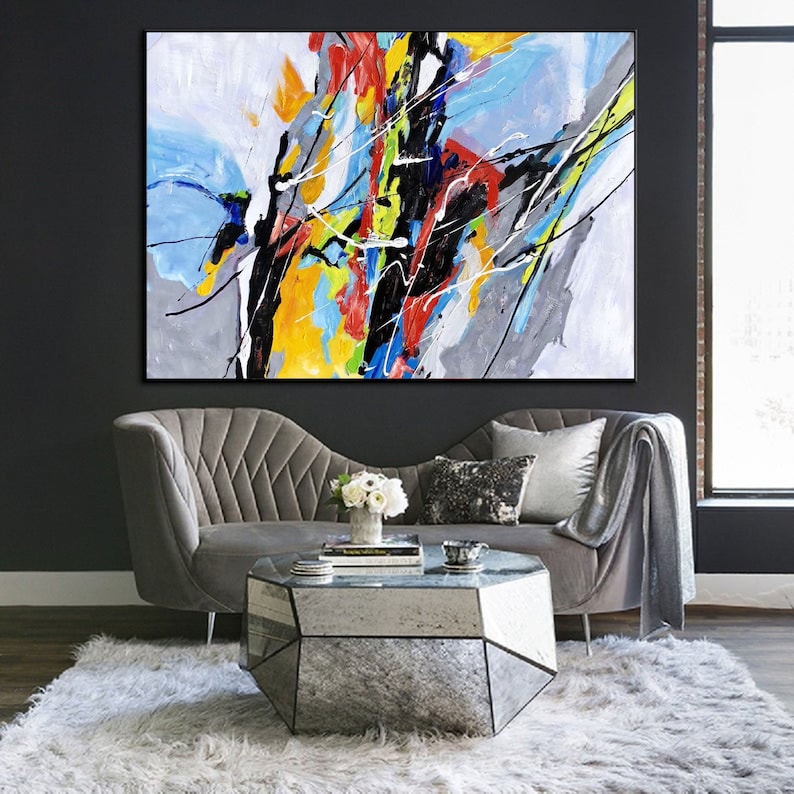 May include: Abstract painting with bold strokes of black, yellow, red, blue, and green on a gray and white background. The painting is hanging above a gray velvet sofa with a mirrored coffee table.