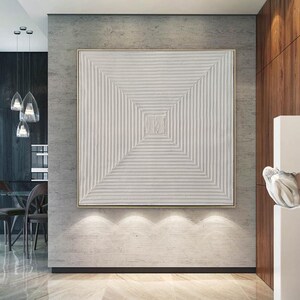 May include: A white textured abstract painting with a geometric design in a gold frame hanging on a gray wall in a modern home.