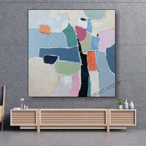 May include: Abstract painting with a black frame. The artwork features a colorful composition of geometric shapes in shades of blue, green, pink, orange, and white. The painting is displayed above a wooden cabinet.
