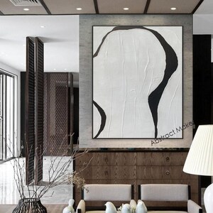 May include: Large abstract painting with a black and white design. The artwork features a textured white background with bold, curved black shapes. The painting is framed and displayed in a modern interior setting.