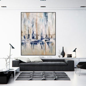 May include: Large abstract painting in a black frame. The artwork features vertical brushstrokes in shades of white, beige, blue, and brown, creating a textured effect. The painting is displayed above a gray sofa in a modern living room setting.