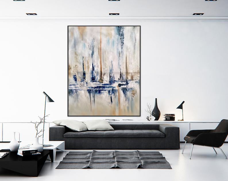 May include: Large abstract painting in a black frame. The artwork features vertical brushstrokes in shades of white, beige, blue, and brown, creating a textured effect. The painting is displayed above a gray sofa in a modern living room setting.