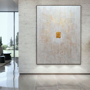 May include: A large rectangular wall art piece featuring a textured white surface with intricate, raised linear patterns and subtle gold accents. A small, square, metallic gold element is centred on the canvas, framed in a thin black border.
