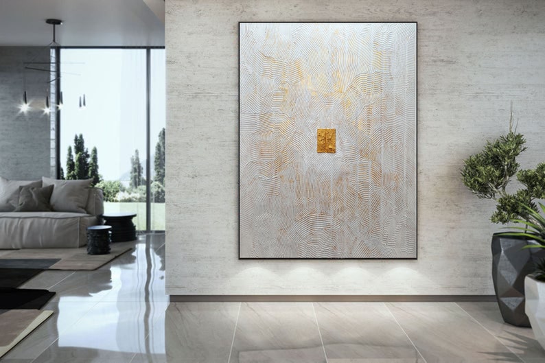 May include: A large rectangular wall art piece featuring a textured white surface with intricate, raised linear patterns and subtle gold accents. A small, square, metallic gold element is centred on the canvas, framed in a thin black border.