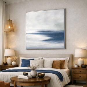 May include: A bedroom scene featuring a large square abstract painting with shades of blue and white, framed in silver. The bed is made with white linens, blue and brown pillows, and a blue throw. Two wooden nightstands with lamps flank the bed.