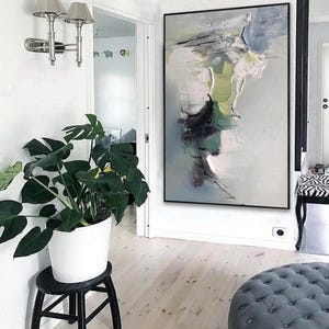 May include: Abstract painting in shades of green, white, and gray with black accents. The painting is framed in a black frame and hangs on a white wall in a hallway.