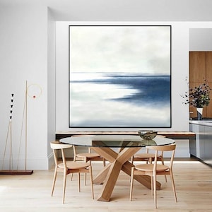 May include: A large abstract painting with a black frame dominates a dining room. The artwork features shades of blue and white, evoking a seascape. A glass-topped table with wooden chairs and a decorative floor lamp complete the room.