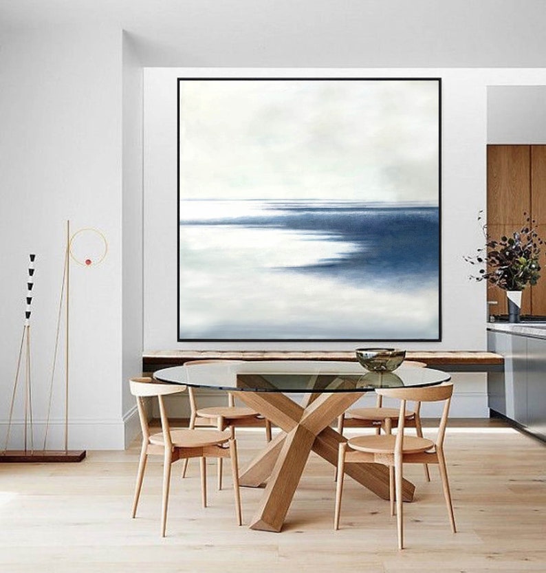 May include: A large abstract painting with a black frame dominates a dining room. The artwork features shades of blue and white, evoking a seascape. A glass-topped table with wooden chairs and a decorative floor lamp complete the room.