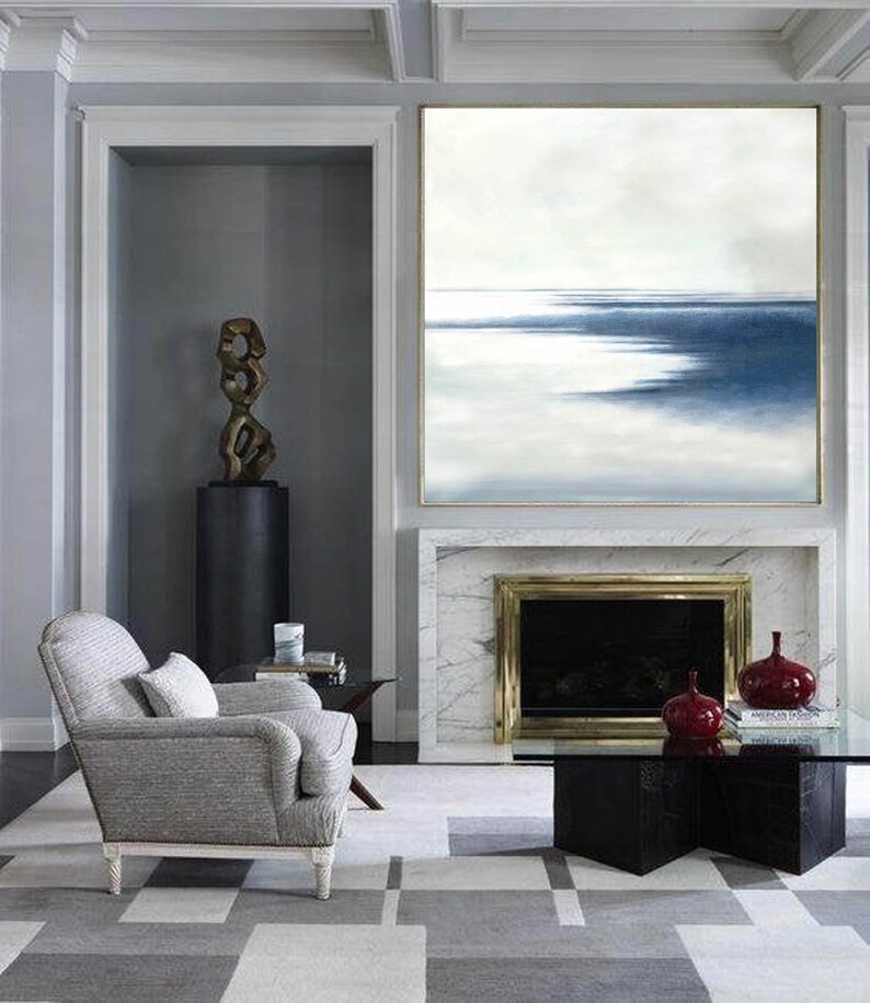 May include: An interior living room scene featuring a large abstract painting with blue and white hues above a fireplace. A gray armchair, a black coffee table, and a geometric patterned rug complete the space. Decorative red vases sit on the table.