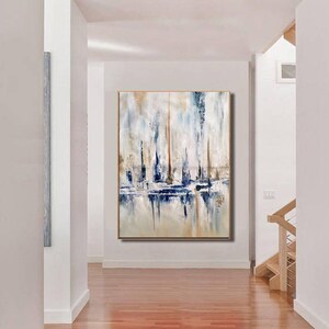 May include: Abstract painting with a gold frame. The artwork features vertical brushstrokes in shades of blue, white, and tan, creating a textured effect. The painting is displayed in a hallway with hardwood floors and white walls.