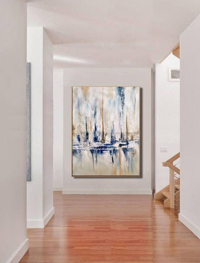 May include: Abstract painting with a gold frame. The artwork features vertical brushstrokes in shades of blue, white, and tan, creating a textured effect. The painting is displayed in a hallway with hardwood floors and white walls.