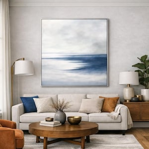 May include: A large framed abstract painting with a seascape theme dominates a living room. The artwork features shades of blue and white, evoking a serene ocean scene. The room includes a white sofa, pillows, a wooden coffee table, and a floor lamp.