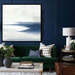 May include: An abstract painting with a black frame, depicting a seascape in shades of blue and white. The artwork hangs above a green velvet sofa with decorative pillows. A white lamp and side table are also visible.