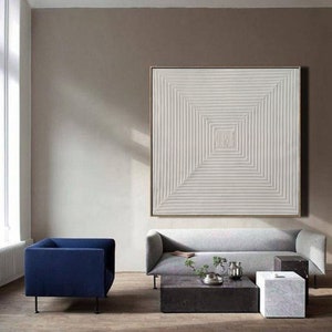 May include: A white abstract painting with a geometric pattern of lines creating a square in the center. The painting is hanging on a wall in a living room with a gray sofa and a blue armchair.