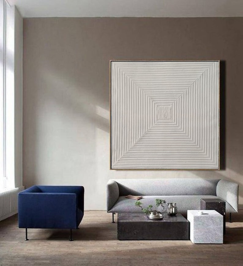 May include: A white abstract painting with a geometric pattern of lines creating a square in the center. The painting is hanging on a wall in a living room with a gray sofa and a blue armchair.