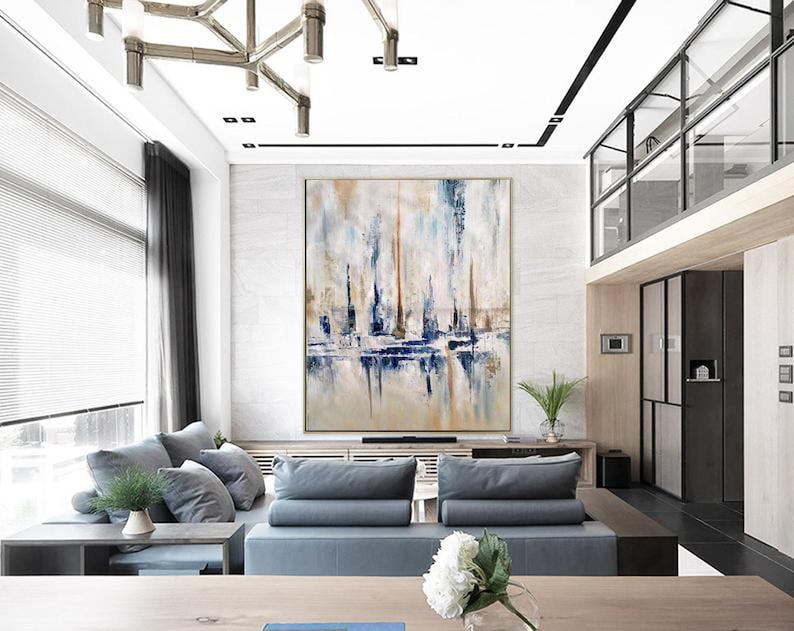 May include: Large abstract painting with vertical brushstrokes in shades of blue, beige, and white. The artwork is displayed in a modern living room with gray sofas, a wooden coffee table, and a decorative chandelier.