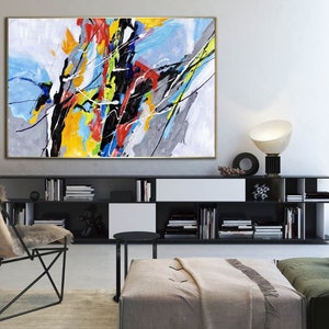 May include: Abstract painting with bold strokes of yellow, red, blue, black, and white on a canvas. The painting is framed in gold and hangs on a white wall above a black bookshelf.