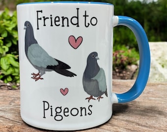 May include: White ceramic mug with a blue handle and rim, featuring the text "Friend to Pigeons" and illustrations of two pigeons and pink hearts. The mug is designed for hot drinks.