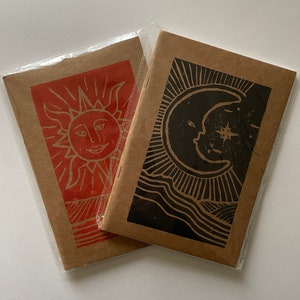 Puede incluir: Dos pequeños cuadernos de papel kraft con un diseño de sol y luna. El cuaderno del sol es rojo y el cuaderno de la luna es negro.
