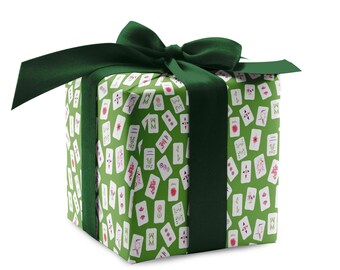 May include: A square gift box wrapped in green paper with a pattern of white and red playing card illustrations. The box is tied with a dark green satin ribbon and bow, creating a festive presentation.