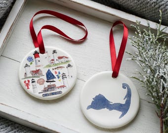 May include: Two white ceramic ornaments with red ribbon hangers. One features a Cape Cod illustration with lighthouses, a lobster, and the text "CAPE COD." The other has a blue Cape Cod outline.