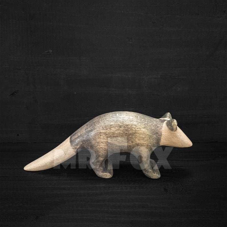 May include: A wooden anteater sculpture with a light brown head and tail, and a gray body. The sculpture is carved with visible wood grain and stands on four legs. The background is a dark wood surface.