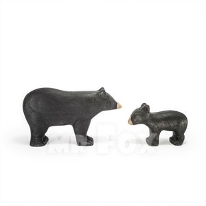 May include: Two wooden bear figurines. One is a larger adult bear, and the other is a smaller cub. Both are painted black with tan-colored snouts. The figurines are standing on a white surface.
