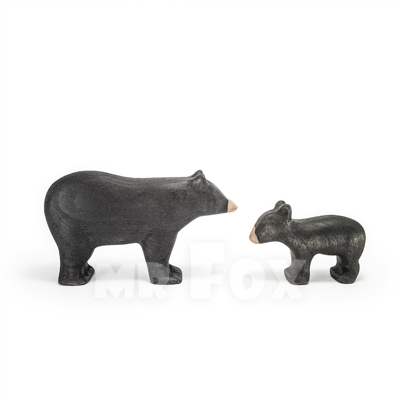 May include: Two wooden bear figurines. One is a larger adult bear, and the other is a smaller cub. Both are painted black with tan-colored snouts. The figurines are standing on a white surface.