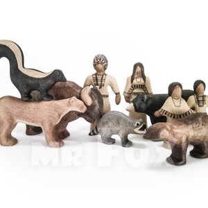 May include: Wooden toy set featuring a skunk, cougar, bison, raccoon, bear, and Native American figures. The animals are in various shades of brown and gray, while the people are in tan and cream colors. The skunk is black and white.