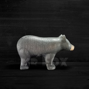 May include: A gray bear figurine with a light brown snout stands on a dark wooden surface. The bear is carved with visible texture, showcasing its form. The figurine is a decorative item.
