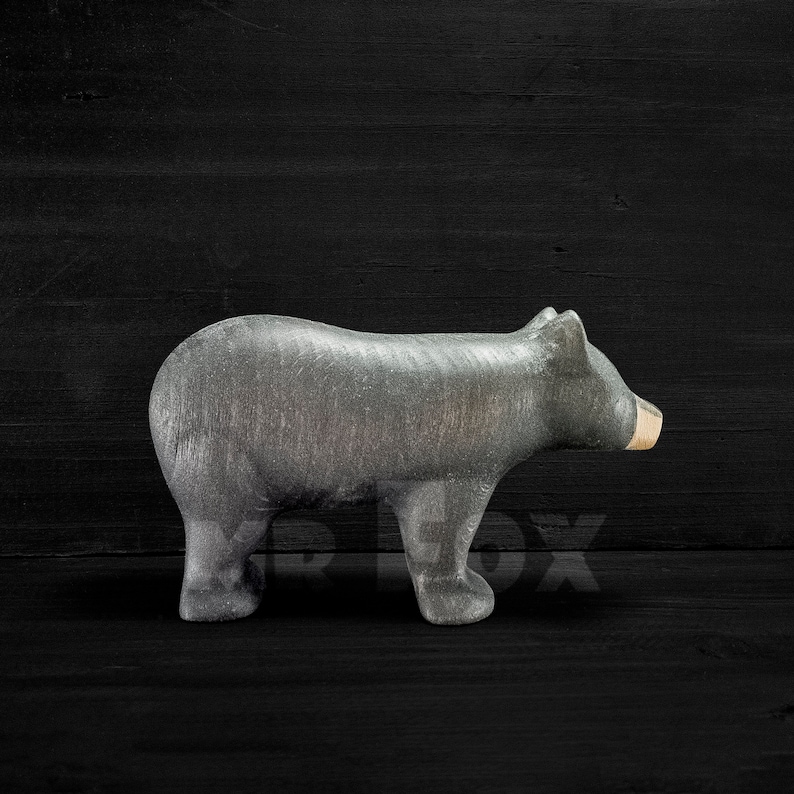 May include: A gray bear figurine with a light brown snout stands on a dark wooden surface. The bear is carved with visible texture, showcasing its form. The figurine is a decorative item.