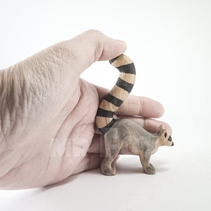 May include: A wooden hand-carved lemur figurine with a black and brown striped tail. The lemur is standing on a white surface.