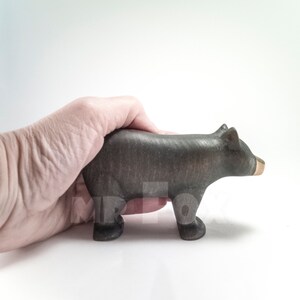 May include: A wooden black bear figurine with a gold-colored snout. The bear is standing on all fours and has a simple, stylized design. The figurine is held in a hand against a white background.
