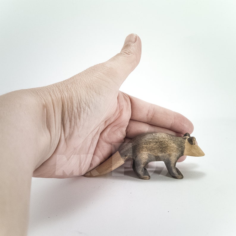 May include: A small, wooden raccoon figurine with a striped tail and a light brown snout. The raccoon is painted in shades of brown and gray. The figurine is held in a hand against a white background.