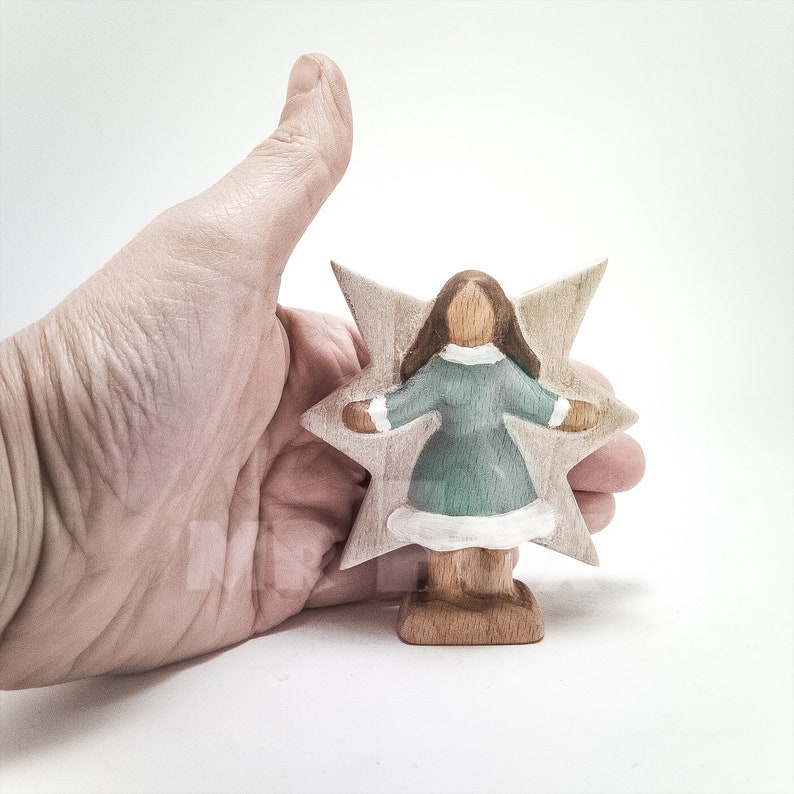 Wooden winter fairy figurine against a female hand for size reference. Front view. White background. Blue dress. White showflake shaped wings. Long dark brown hair.