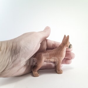 May include: A small, hand-carved wooden dog figurine in a reddish-brown color. The dog is standing on all four legs and has its head turned to the side. The figurine is being held in a person's hand.