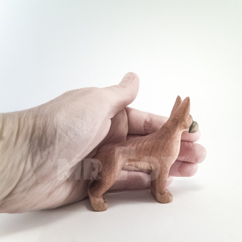 May include: A small, hand-carved wooden dog figurine in a reddish-brown color. The dog is standing on all four legs and has its head turned to the side. The figurine is being held in a person's hand.
