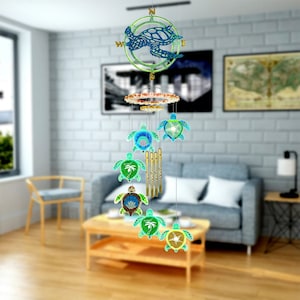 Puede incluir: Una carillón de viento decorativo con un diseño de tortuga marina. La parte superior tiene una tortuga azul dentro de un círculo verde con puntos cardinales. Debajo hay varios carillones con forma de tortuga coloridos y tubos dorados. Los carillones cuelgan de una parte superior decorativa de varias capas.