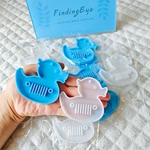 Puede incluir: Cinco moldes de silicona para hacer patitos de goma. Los moldes tienen forma de pato con una superficie texturizada. Dos moldes son blancos y tres son azules.